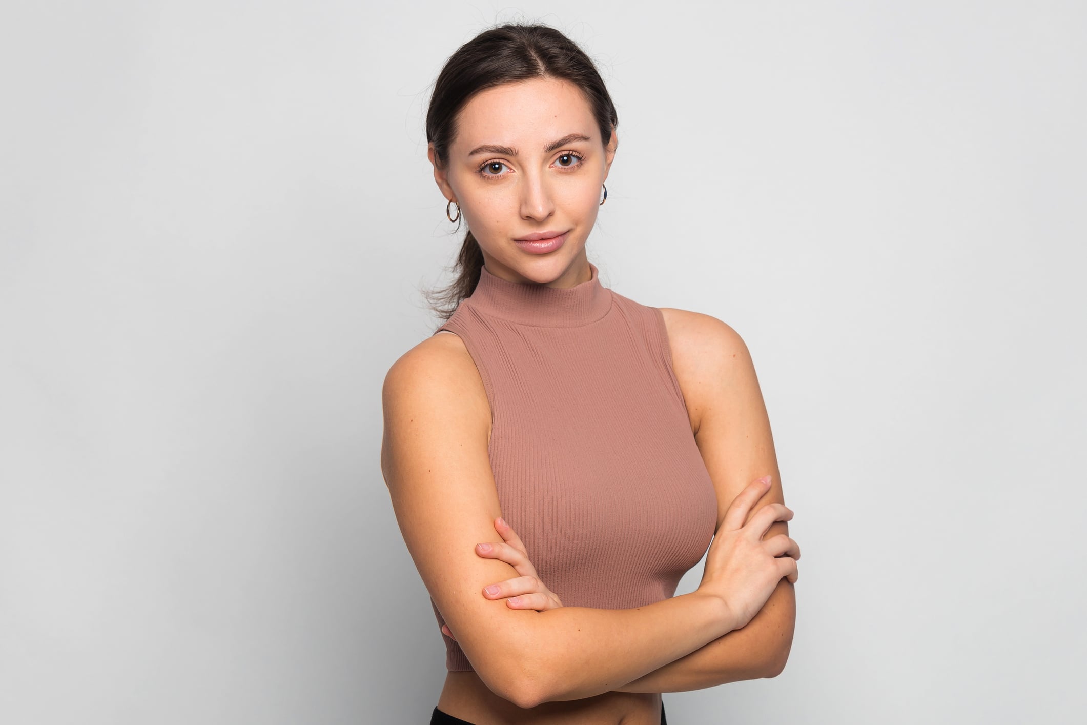 Beautiful young woman portrait with crossed arms. Successful woman looking at camera isolated on gray, copy space for text. Studio shot of good looking female dressed in short turtleneck